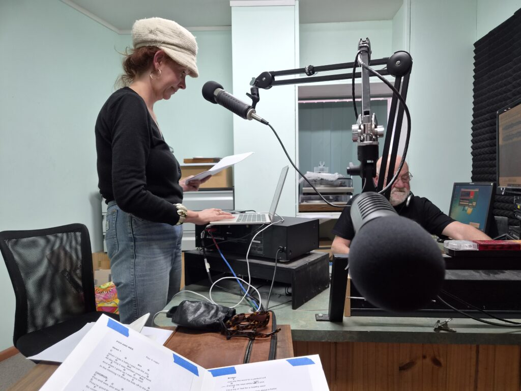 Interior of a radio station. A woman with a jaunty cap stands on the left near a microphone, and a bearded man in the background operates a console.